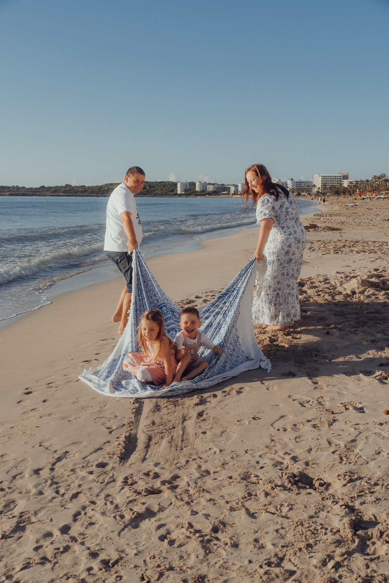 Mutter und Vater ziehen 2 kleine Kinder auf einer großen Decke am Strand von Cala Millor