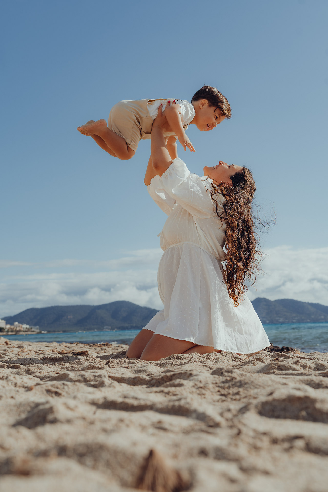 Eine Mutter hält ihren kleinen Sohn am Strand hoch in die Luft und zeigt dabei ihren neuen Babybauch